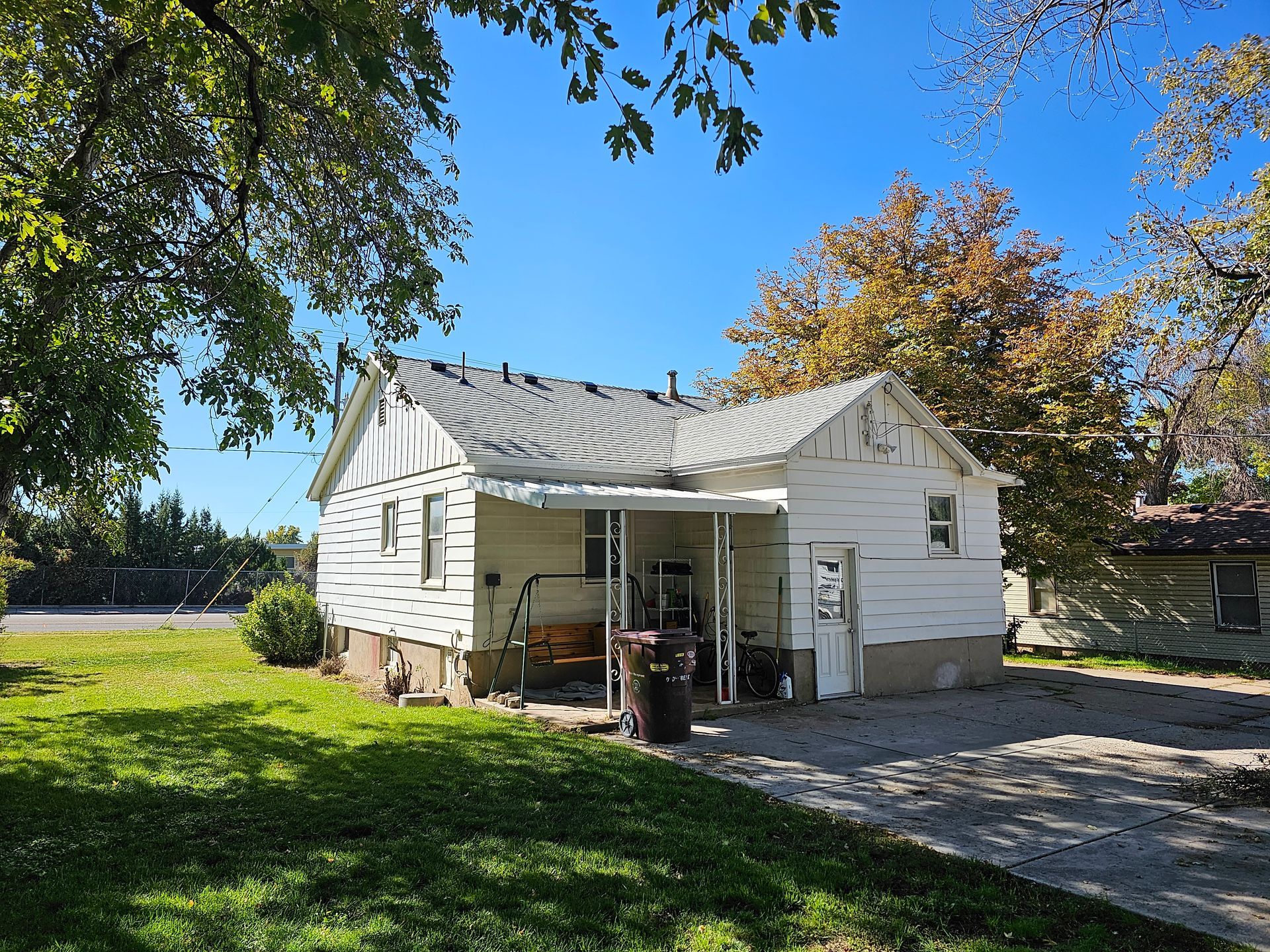 A small white house with a porch and a trash can in front of it.