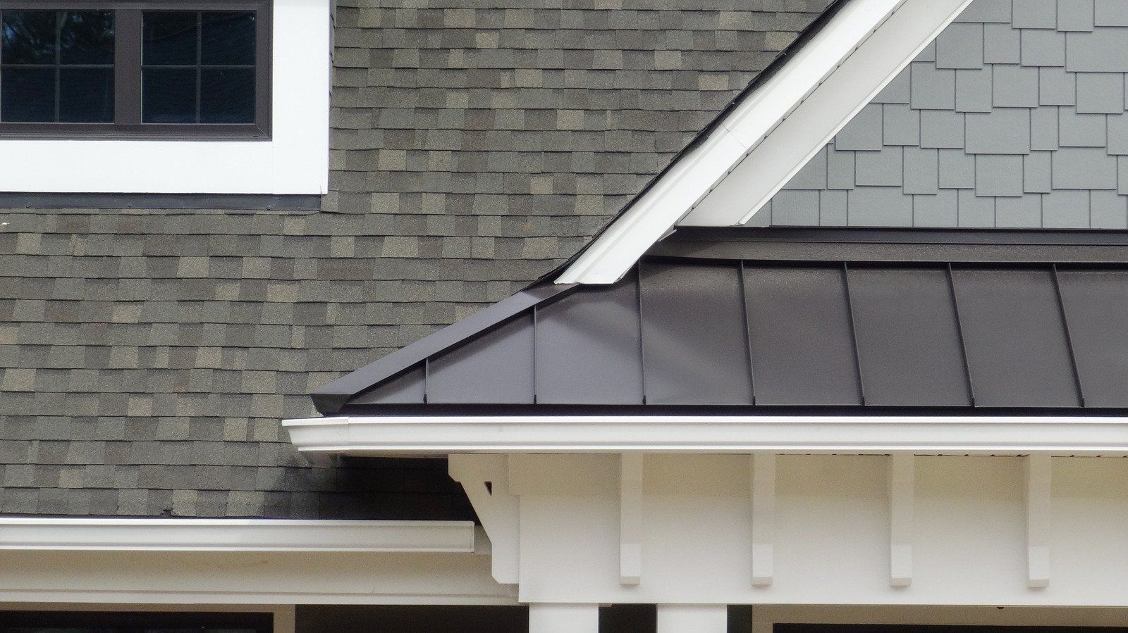 Close-up of a house roof with various materials: shingles, dark metal, and white trim.