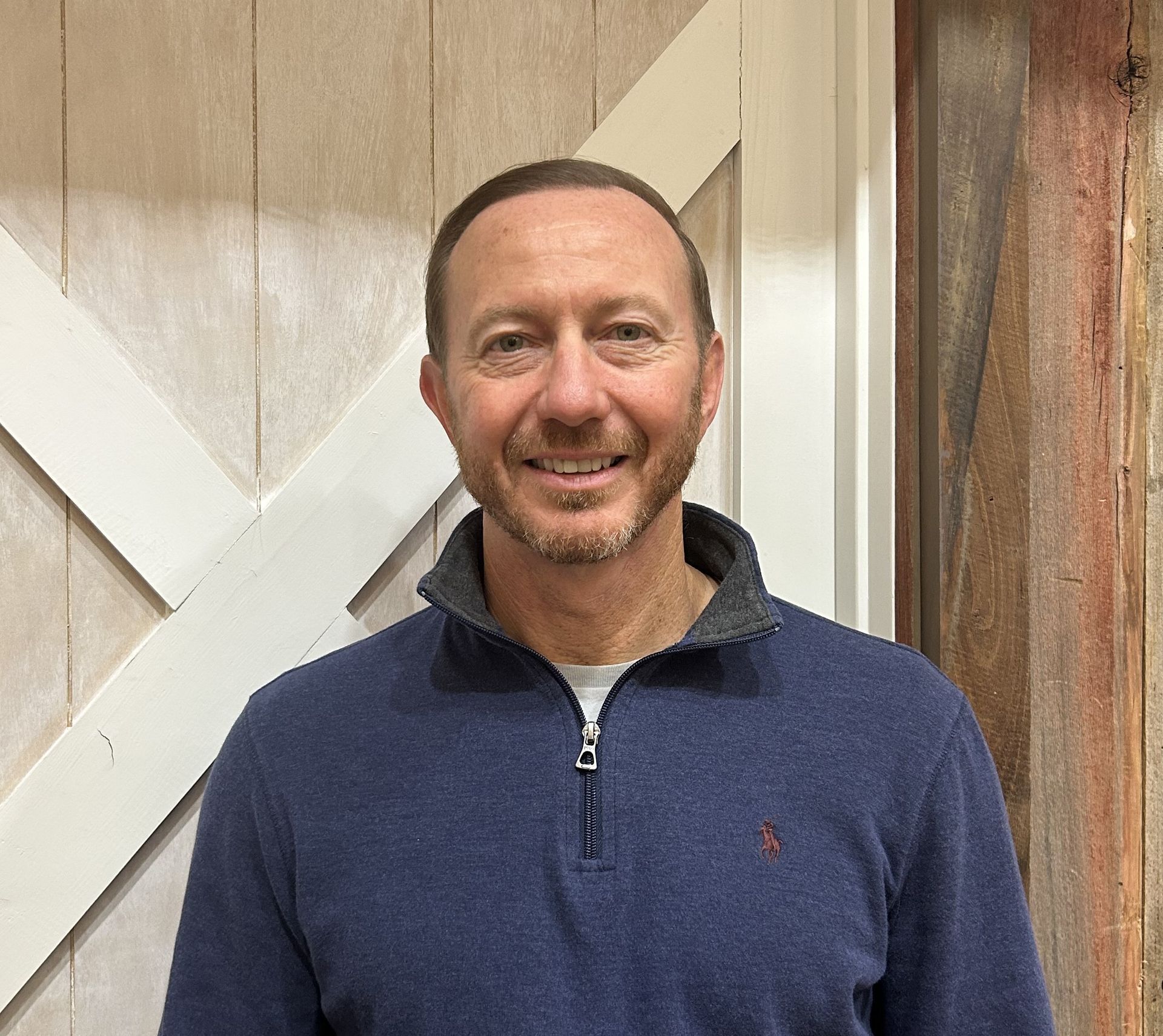 Man with glasses smiles, wearing plaid shirt, in front of a brick wall.