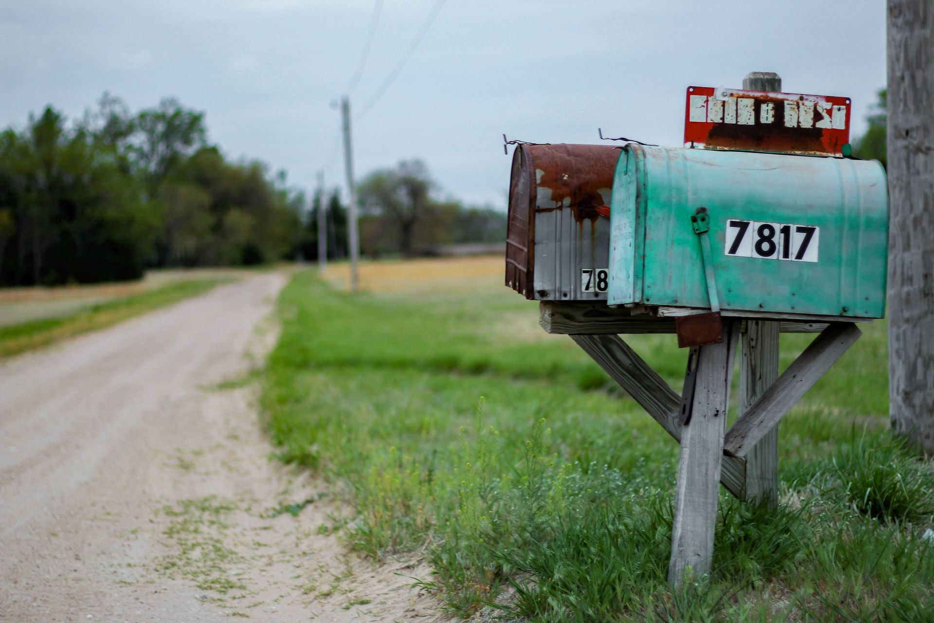 Weathered green and brown mailboxes on a wooden post beside a dirt road with green fields and cloudy sky.