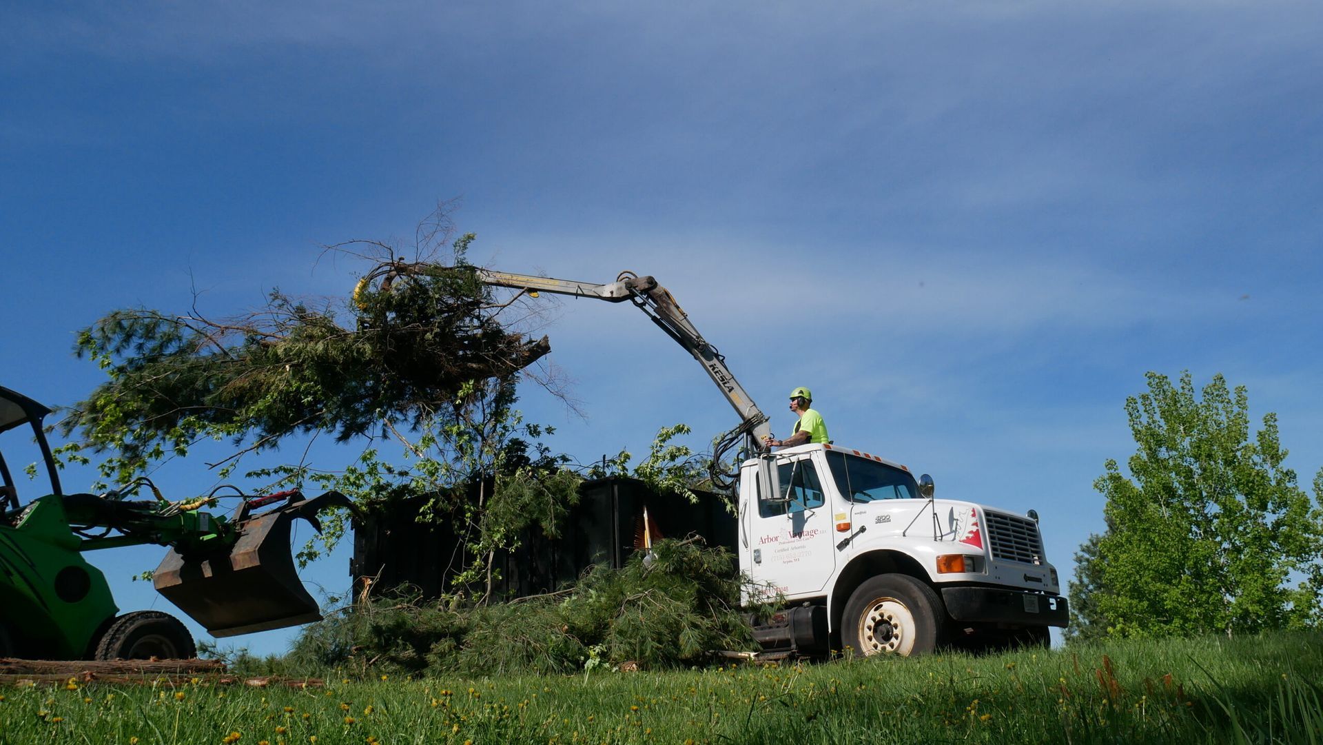 A white truck is being loaded with branches and leaves by a tractor.