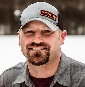 A man with a beard is wearing a stihl hat and smiling.