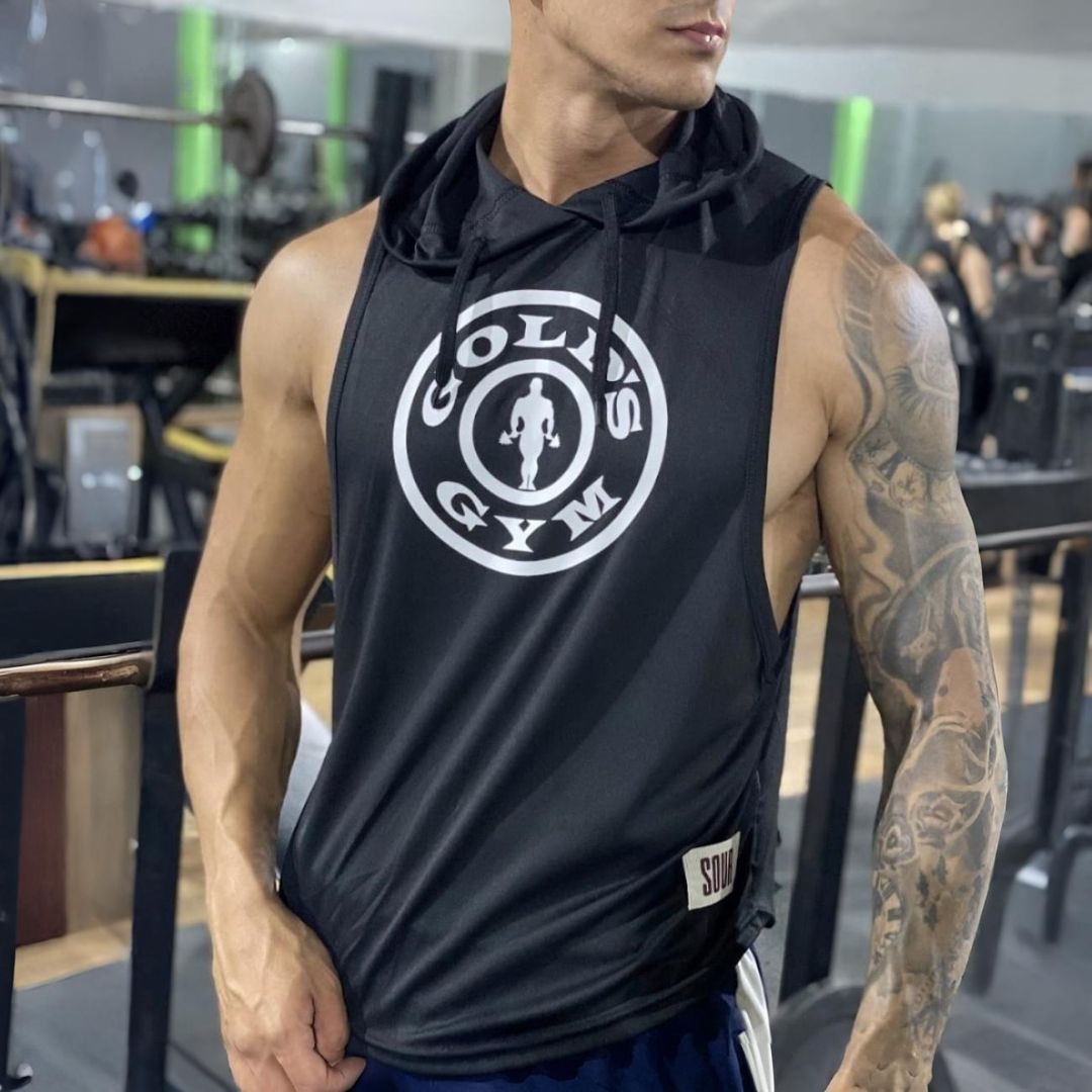 Hombre que viste una camiseta sin mangas con capucha negra de Gold's Gym en un gimnasio, mostrando sus tatuajes en el brazo.
