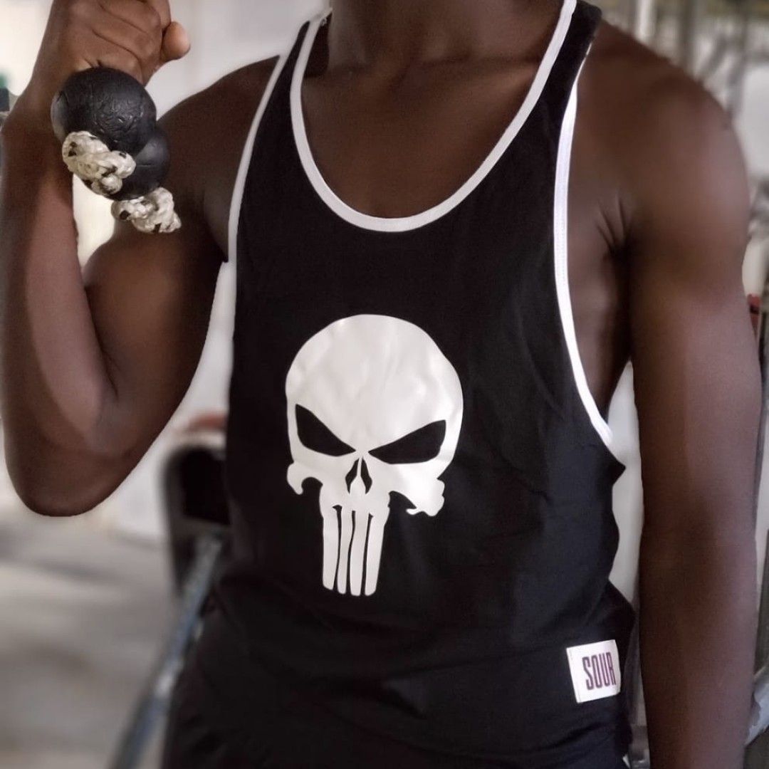 Un hombre con una camiseta negra sin mangas de Punisher flexiona el brazo y sostiene un juguete de perro blanco y negro.