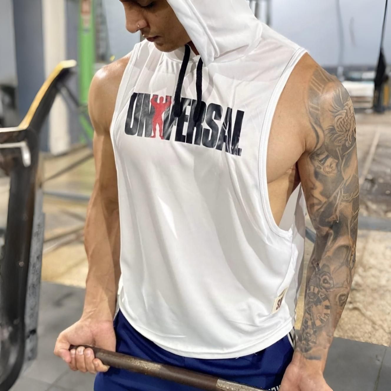Un hombre levanta pesas en un gimnasio. Lleva una sudadera blanca sin mangas con la palabra "UNIVERSAL". Tiene tatuajes.