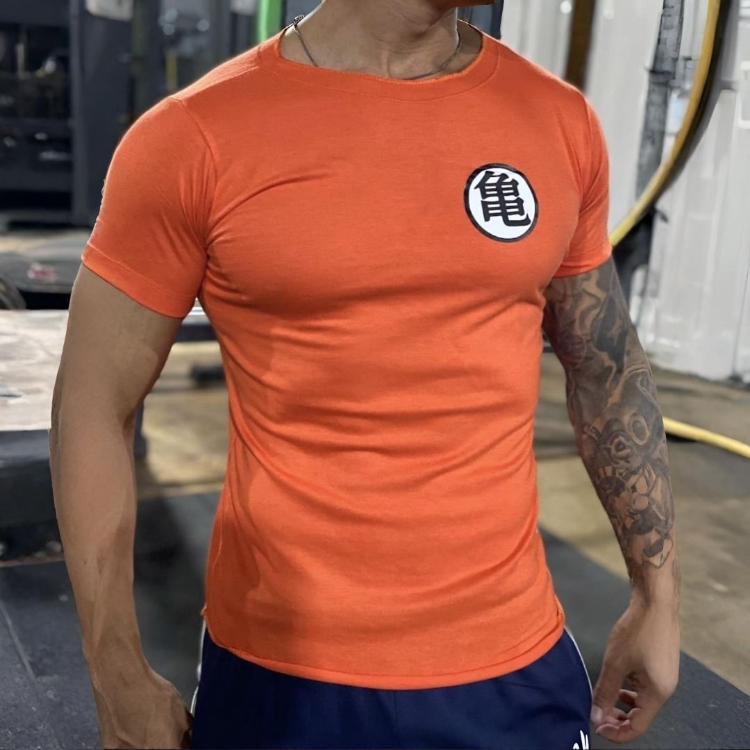 Hombre que lleva una camiseta naranja de Dragon Ball Z con el logo, mostrando brazos musculosos en un gimnasio.