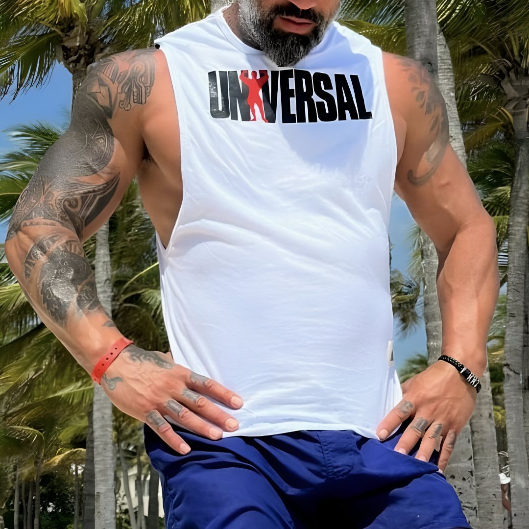 Hombre con tatuajes, vestido con una camiseta blanca y pantalones cortos azules, de pie al aire libre frente a palmeras.