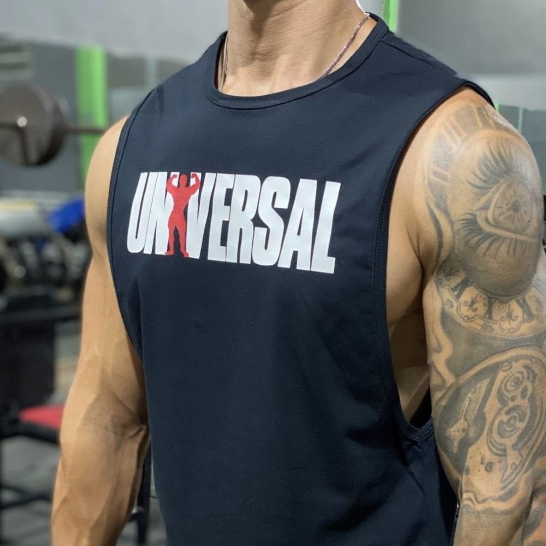 Hombre con una camiseta negra sin mangas "Universal" en un gimnasio, mostrando brazos musculosos con un tatuaje en la manga.