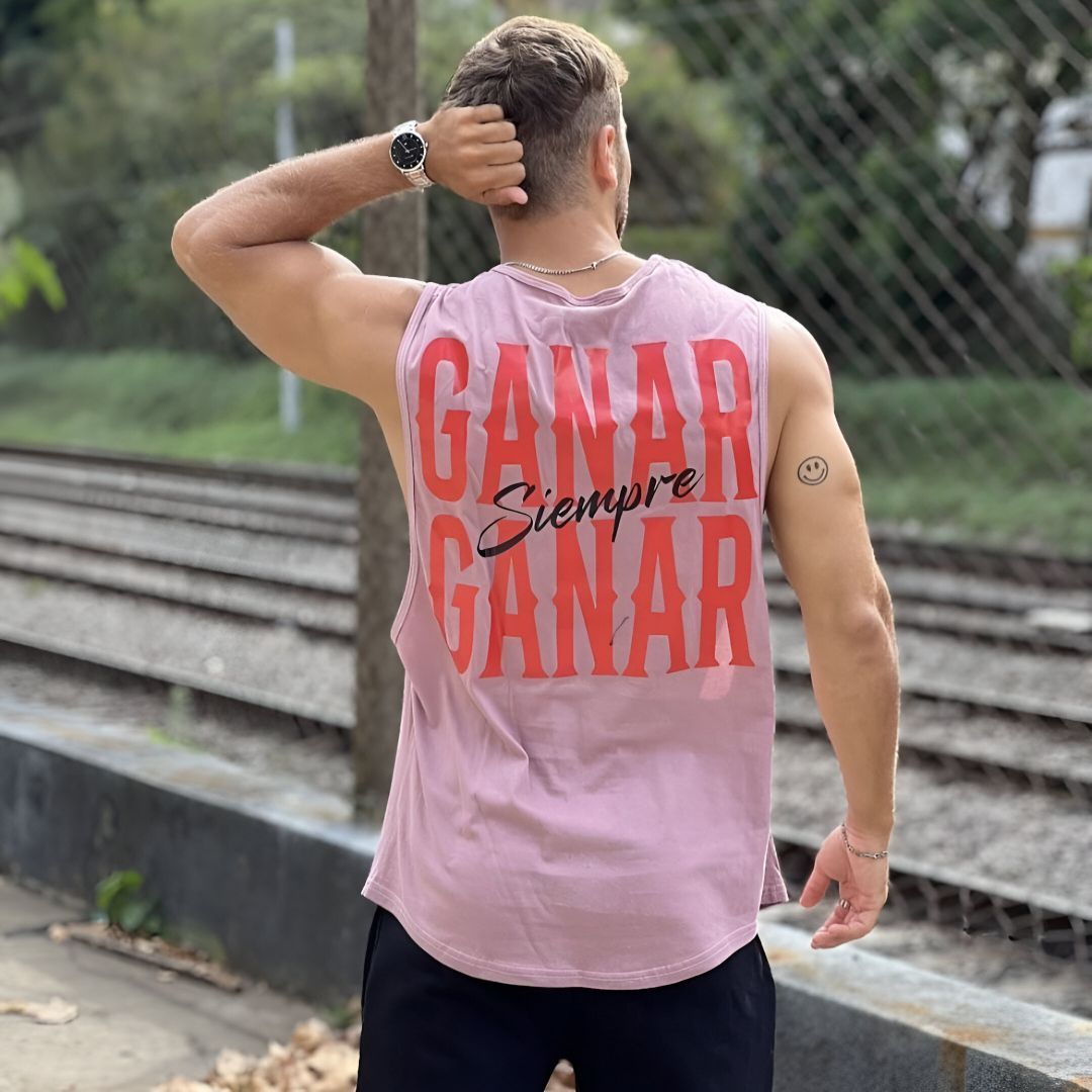 Hombre con una camiseta rosa sin mangas con el texto "GANAR SIEMPRE GANAR", de pie cerca de las vías del tren.