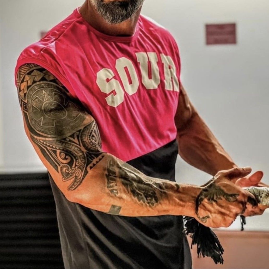 Hombre con tatuajes en el brazo, vestido con una camisa rosa y negra, sosteniendo algo en sus manos.