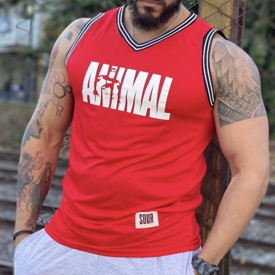 Hombre con camiseta roja con logo “ANIMAL”, tatuajes, apoyado sobre estructura metálica.