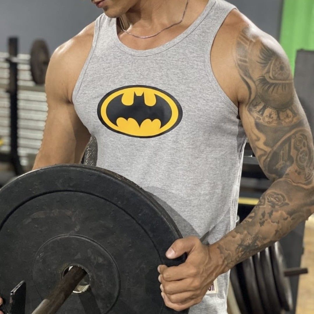 Hombre con camiseta sin mangas de Batman en el gimnasio, sosteniendo pesas y con tatuajes en el brazo.