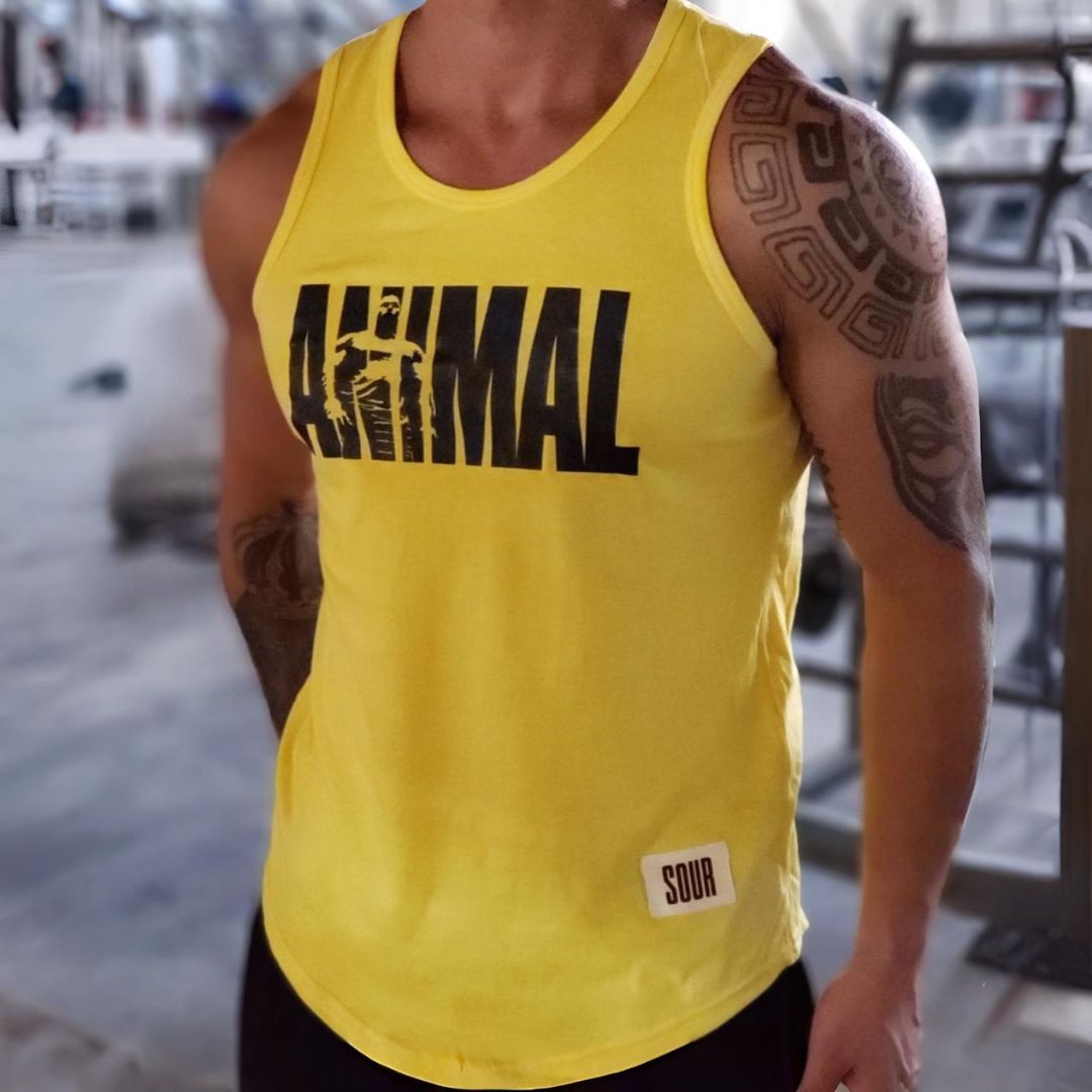 Hombre que lleva una camiseta amarilla sin mangas con la palabra "Animal" en un gimnasio, con tatuajes en el brazo.