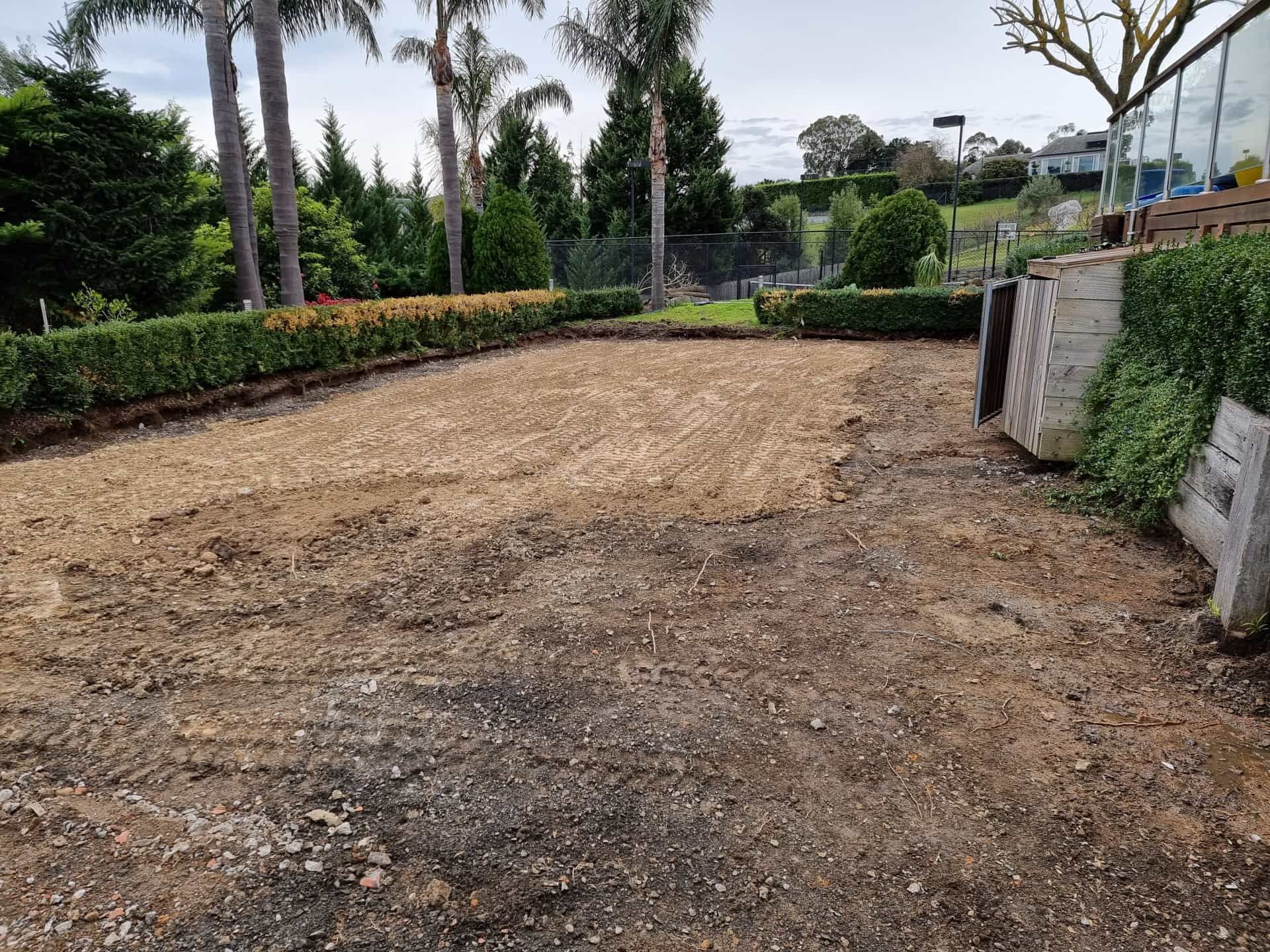 A cleared, flat patch of dirt in a backyard with trimmed hedges, palm trees, and a stone retaining wall in the background  — All Over Pool Removals in Upwey, VIC