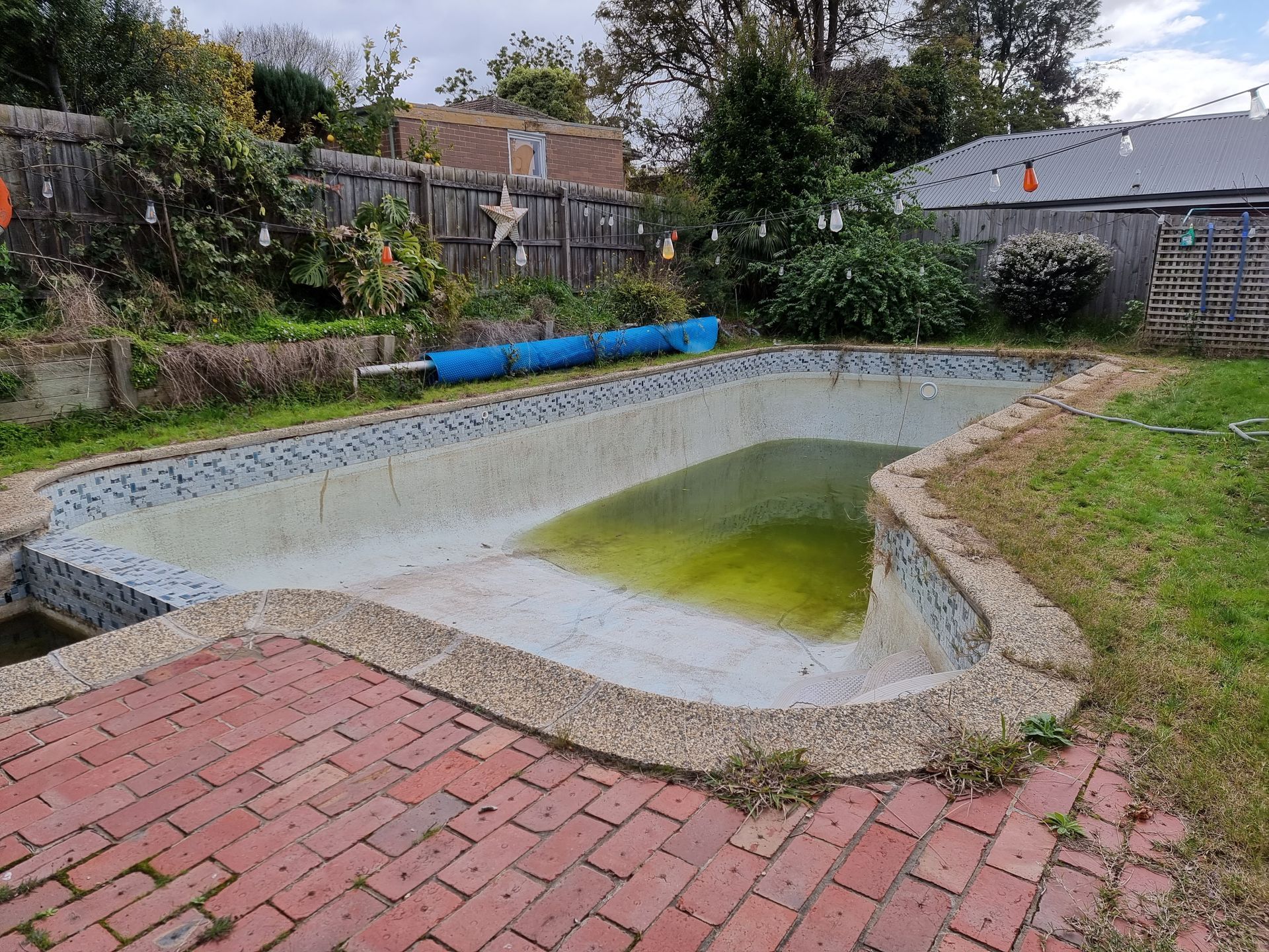 An empty, tiled backyard pool with green stagnant water at the deep end, surrounded by brick paving and a wooden fence  — All Over Pool Removals in Chadstone, VIC