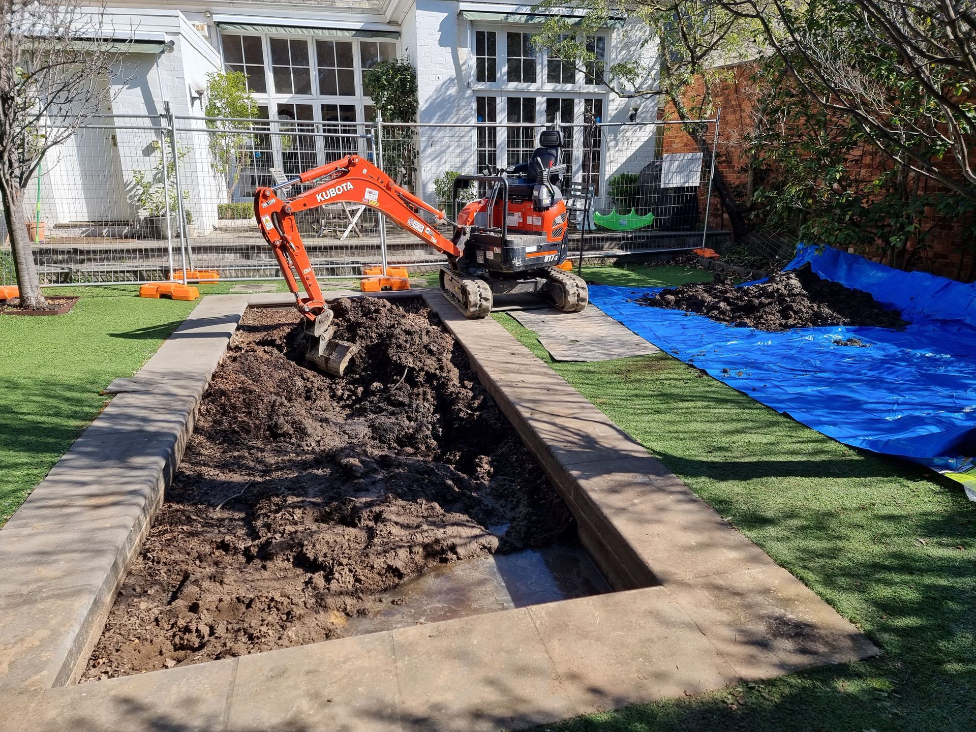 An orange excavator digs into a rectangular trench in a grassy backyard near a house, with soil piled on a blue tarp — All Over Pool Removals in Surrey Hills, VIC