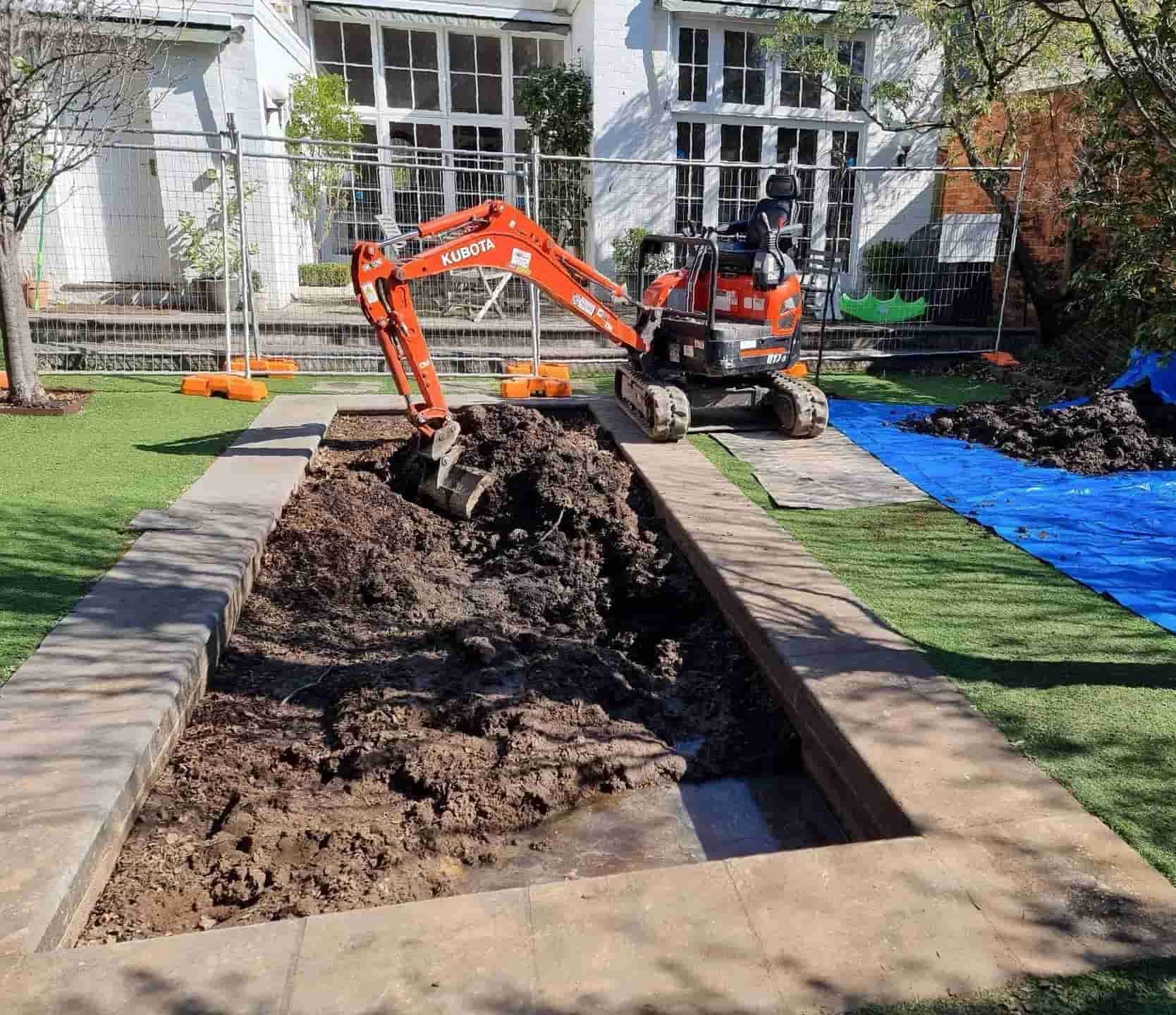 An orange mini excavator digging in a rectangular patch of dirt framed by pavers in a residential backyard — All Over Pool Removals in Glen Waverley, VIC