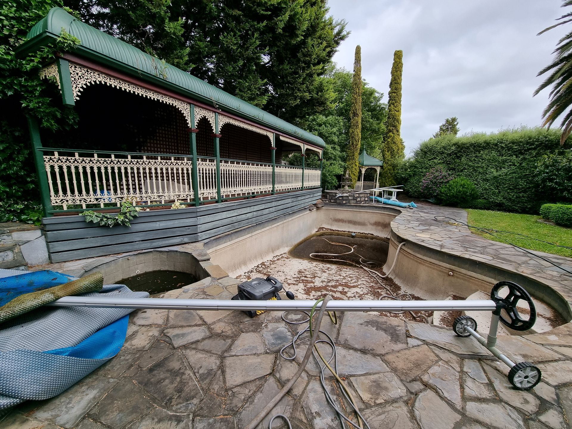A vacant, debris-filled swimming pool next to a stone-paved patio and a decorative, covered veranda — All Over Pool Removals in Hawthorn, VIC