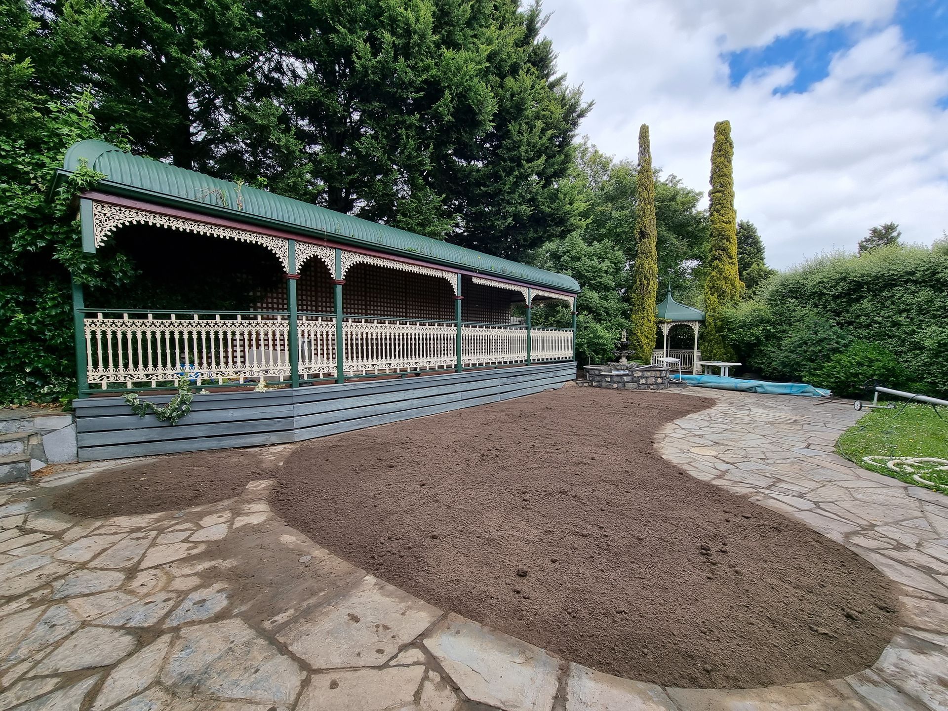 A covered pavilion with a white fence and decorative trim sits beside a large area of dark mulch in a stone-paved garden — All Over Pool Removals in Chadstone, VIC