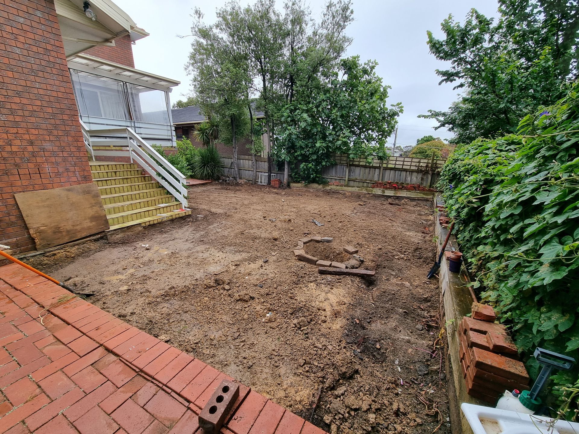 A backyard with bare earth, a brick patio, yellow stairs leading to a porch, and a lush green hedge along the side  — All Over Pool Removals in Templestowe, VIC