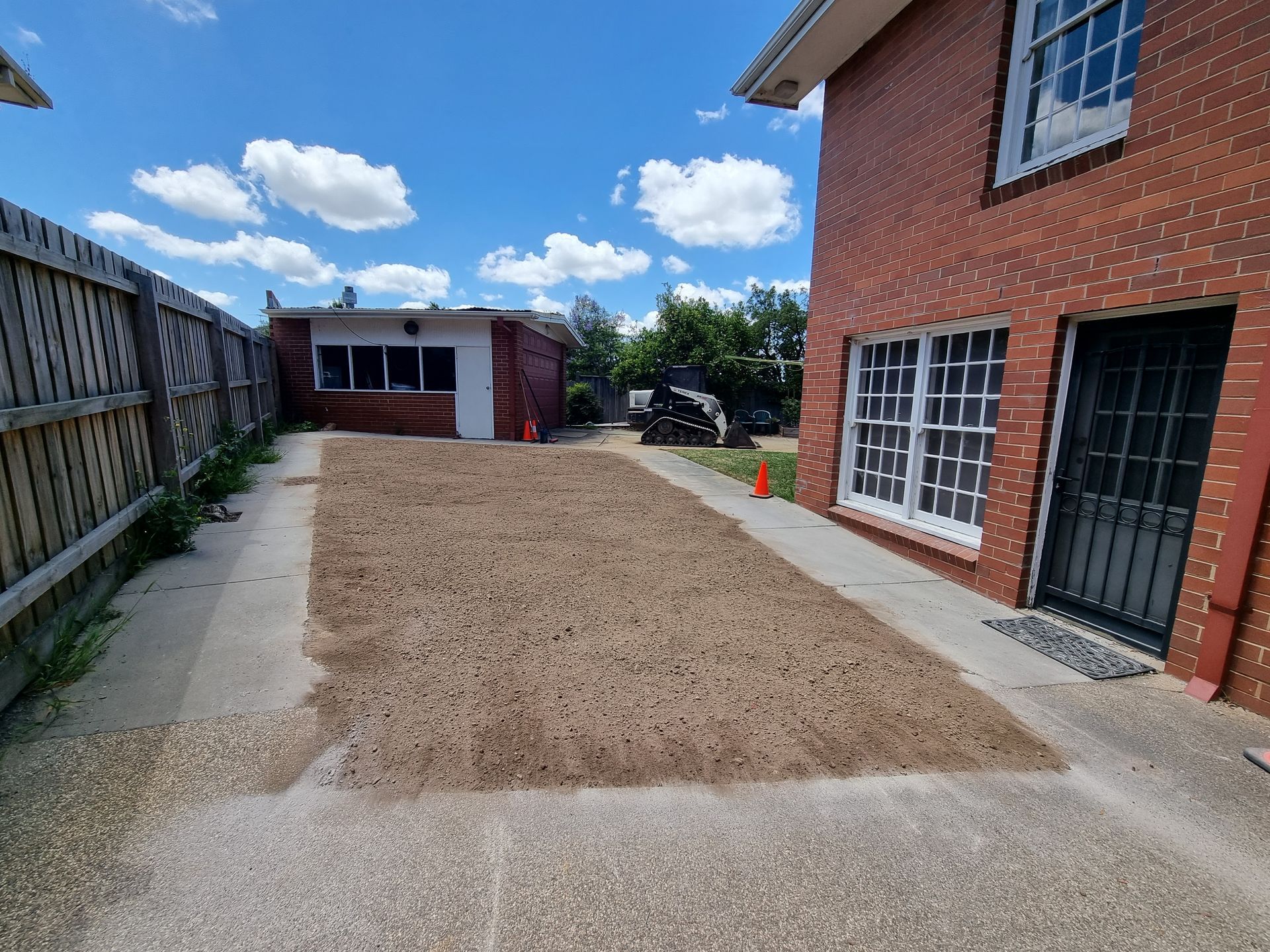 A rectangular patch of dirt sits between a brick house, a wooden fence, and a distant small building under a blue sky — All Over Pool Removals in Surrey Hills, VIC