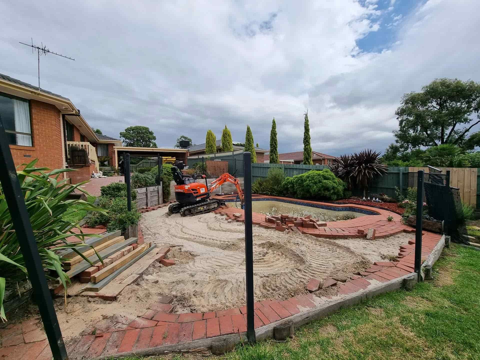 An orange excavator sits in a backyard undergoing construction, surrounded by sandy ground and red brick edging — All Over Pool Removals in Lilydale, VIC