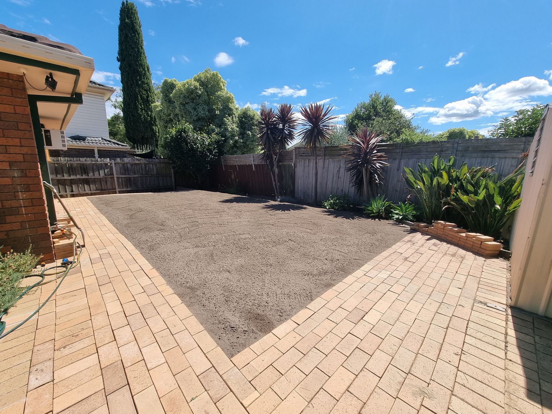 A backyard with a central gravel area framed by light brown brick pavers, a brick wall, and tall foliage under a blue sky — All Over Pool Removals in Surrey Hills, VIC