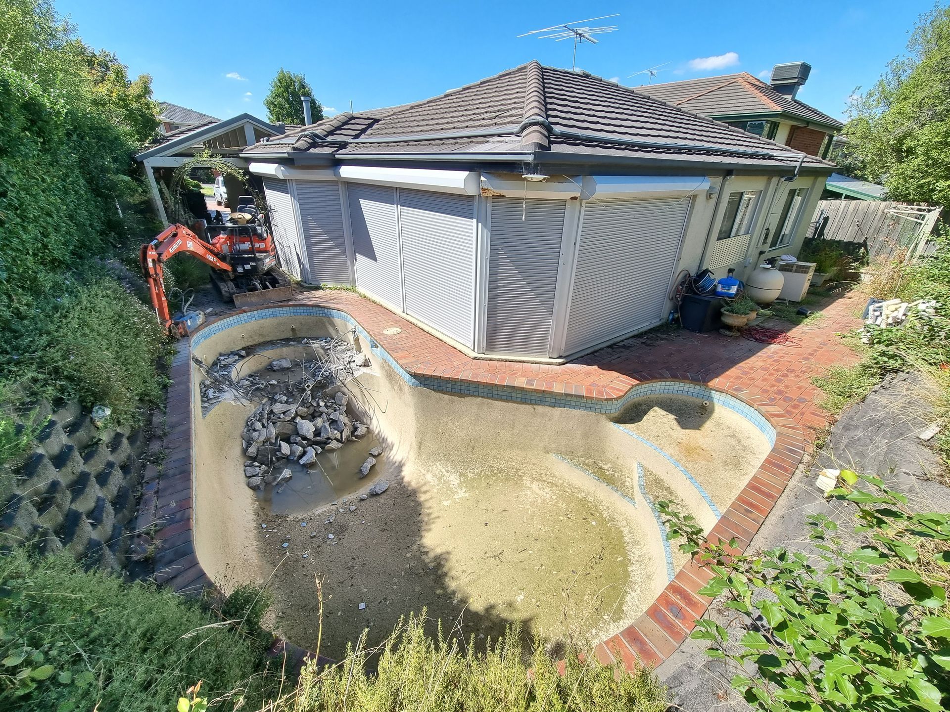 An empty, debris-filled swimming pool under renovation next to a house with closed external shutters on a sunny day  — All Over Pool Removals in Chadstone, VIC