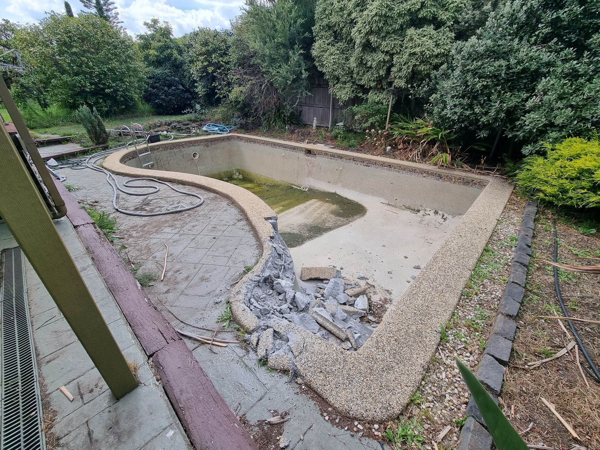 An empty, in-ground swimming pool with exposed aggregate coping and surrounding debris in a backyard garden setting  — All Over Pool Removals in Lilydale, VIC
