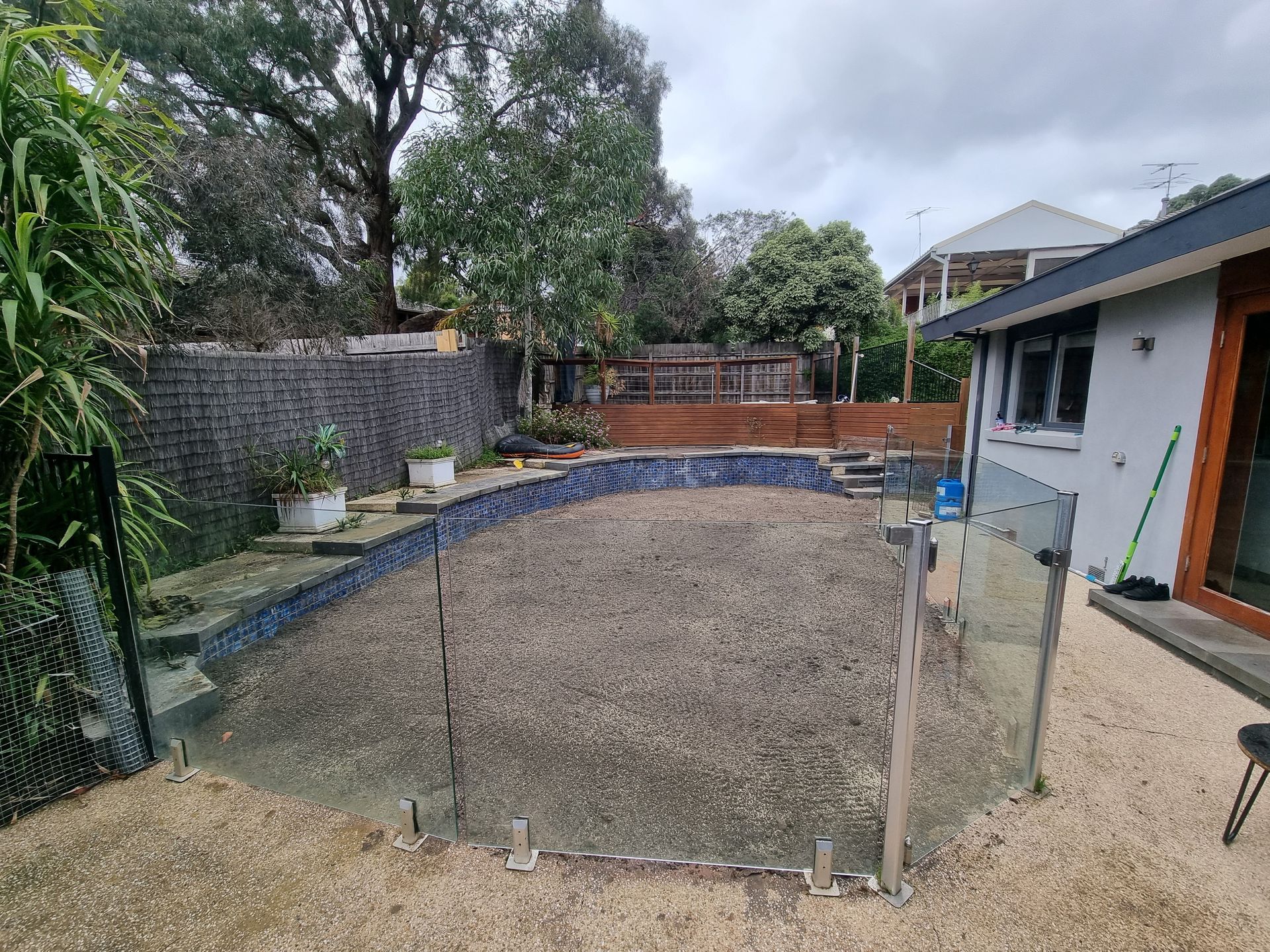An empty, gravel-filled swimming pool surrounded by a glass fence in a backyard next to a house under a cloudy sky  — All Over Pool Removals in Doncaster, VIC