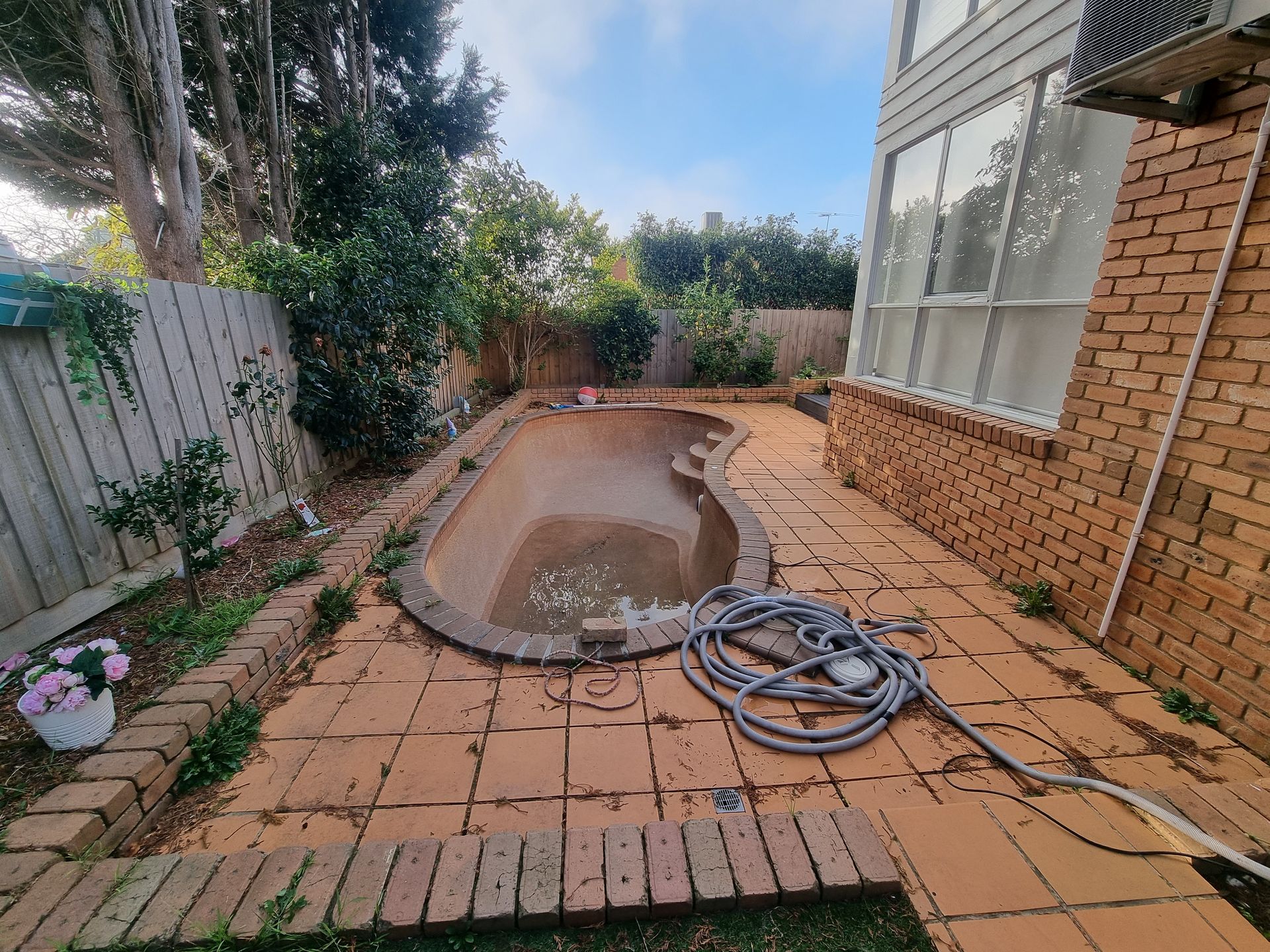 An empty backyard pool with tan paving stones, adjacent to a brick house and wooden fence under a sunny sky — All Over Pool Removals in Beaumaris, VIC