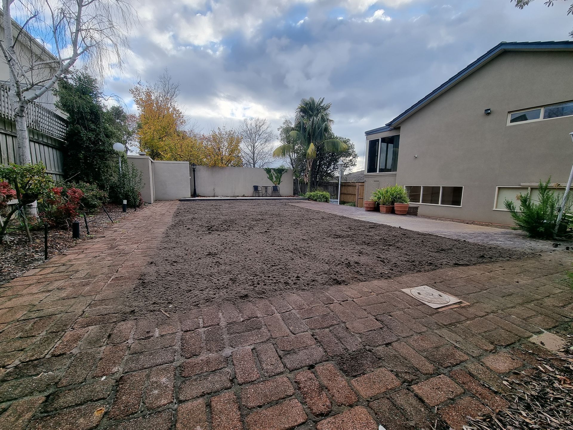 A dirt-covered yard beside a house, viewed from a brick patio under a cloudy sky  — All Over Pool Removals in Doncaster, VIC