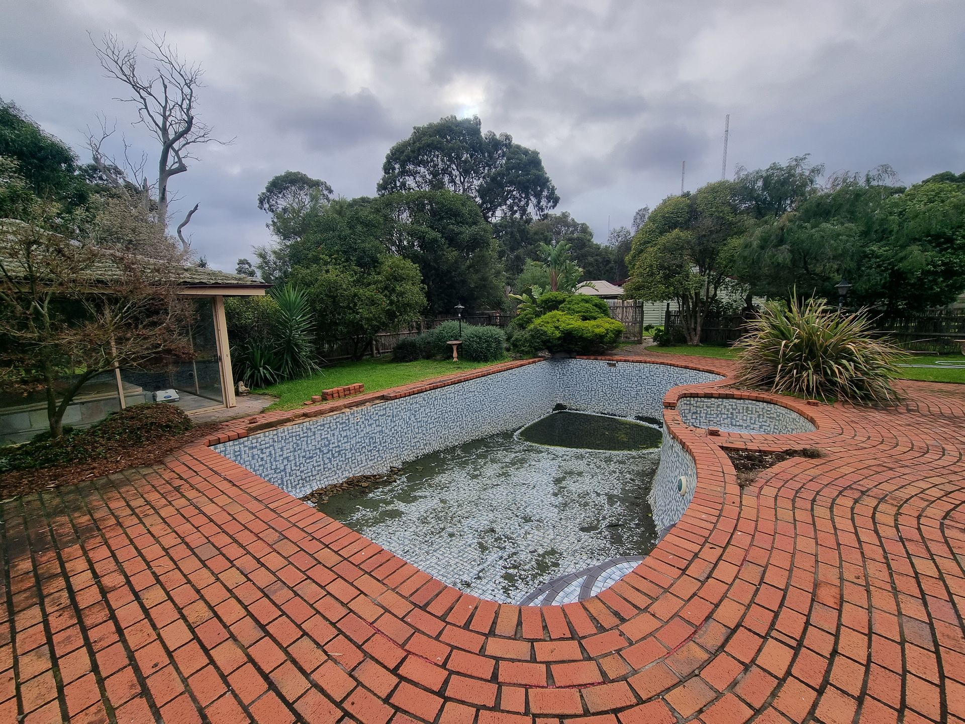 An empty, tiled swimming pool surrounded by a brick patio, set in a backyard garden under a cloudy, overcast sky — All Over Pool Removals in Glen Waverley, VIC