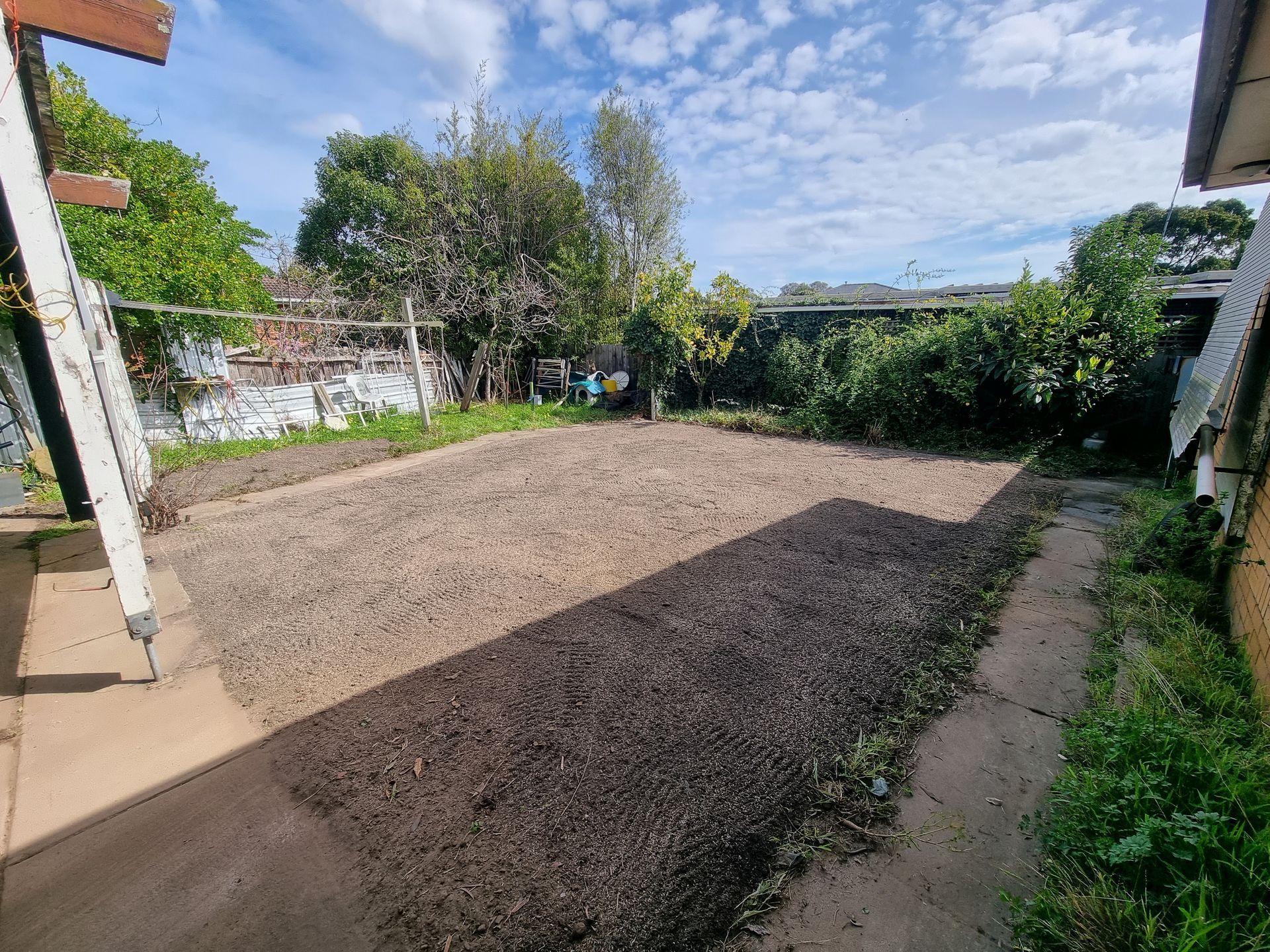 A backyard area featuring a rectangular dirt patch surrounded by mulch, with surrounding trees and a bright, cloudy sky — All Over Pool Removals in Glen Waverley, VIC