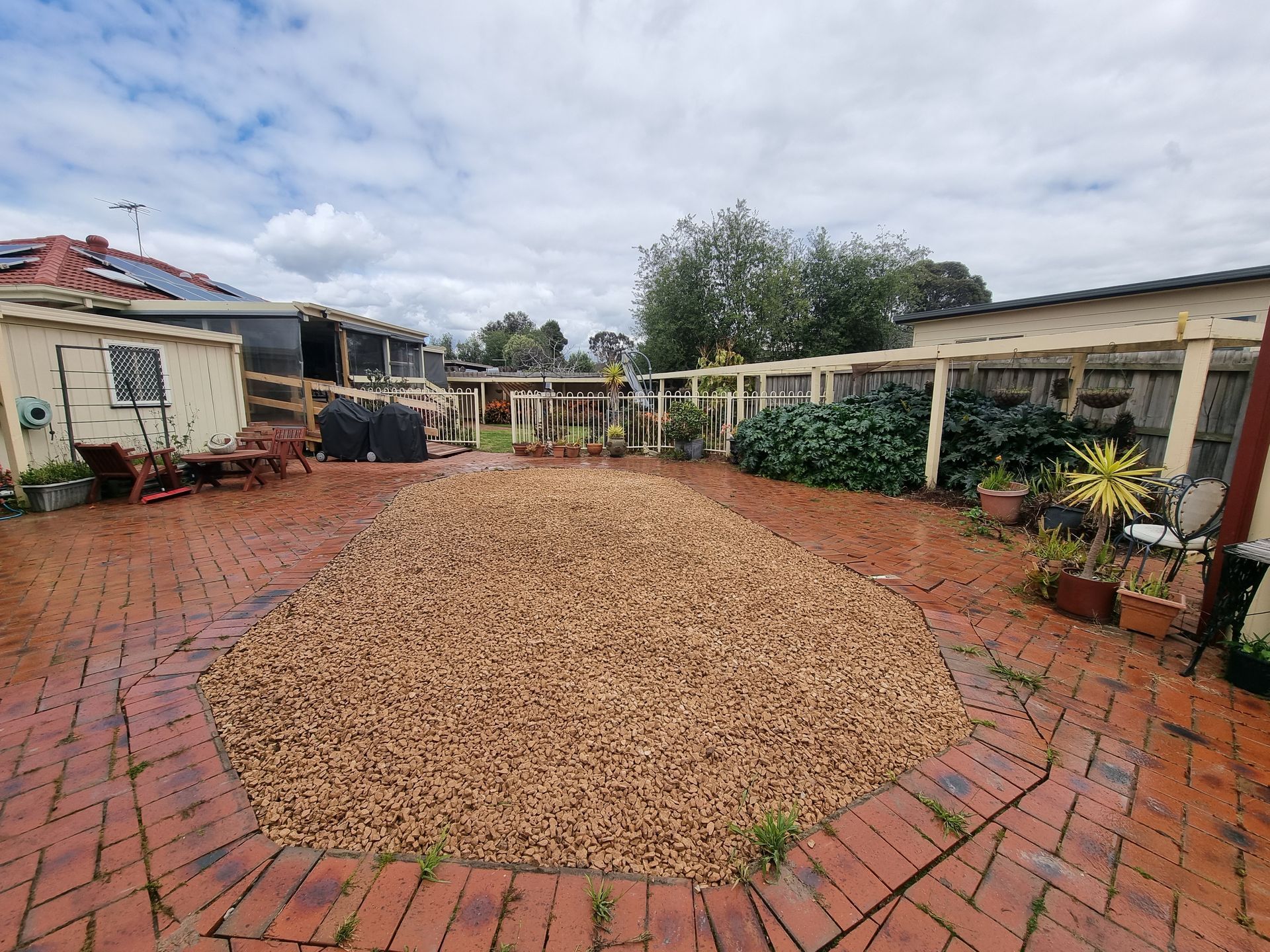 A brick-paved backyard featuring a large central patch of brown gravel, bordered by a wooden trellis and lush greenery  — All Over Pool Removals in Donvale, VIC