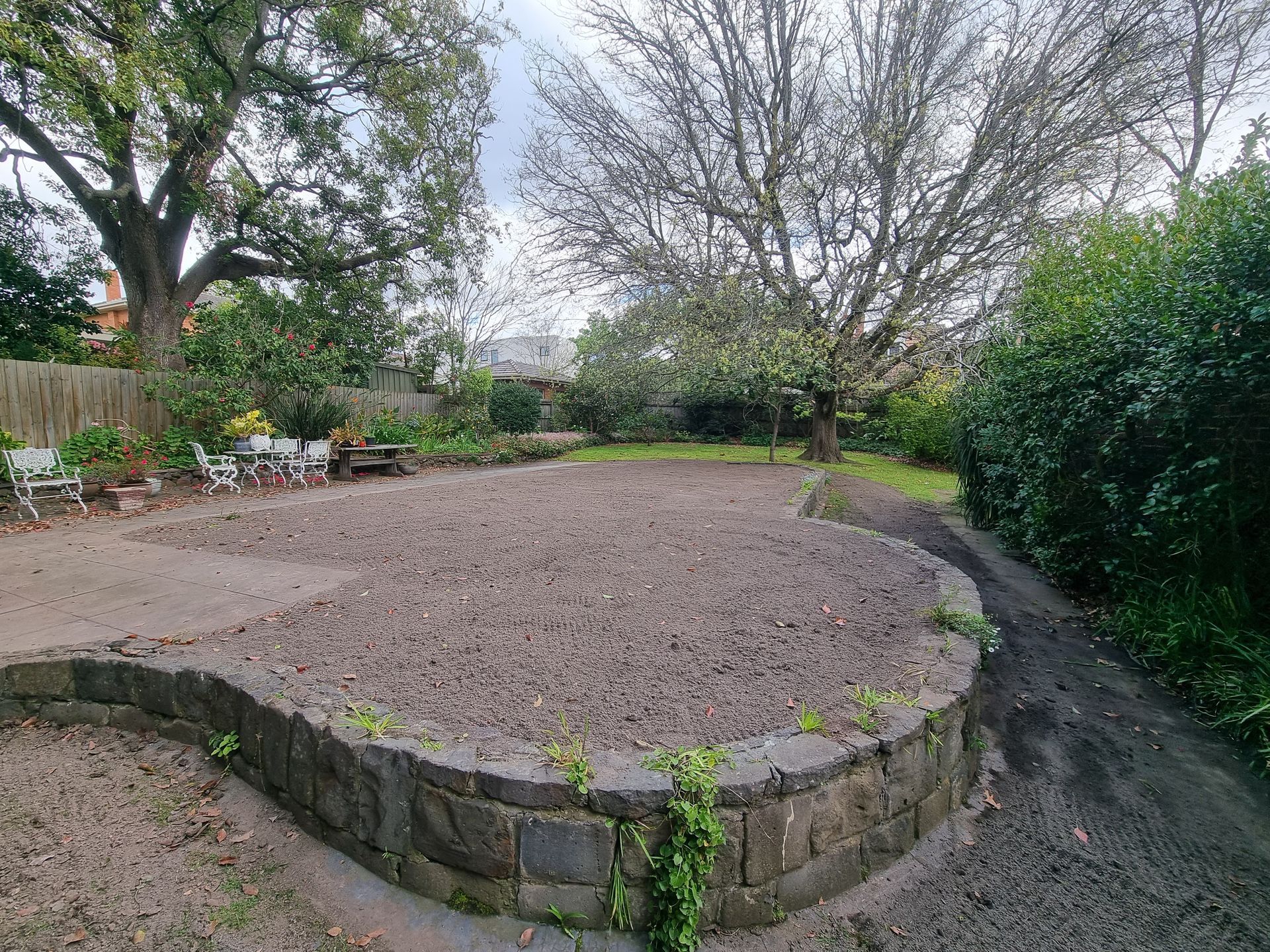 A raised garden bed with a curved stone retaining wall filled with gravel, set in a backyard with trees and shrubbery — All Over Pool Removals in Beaumaris, VIC