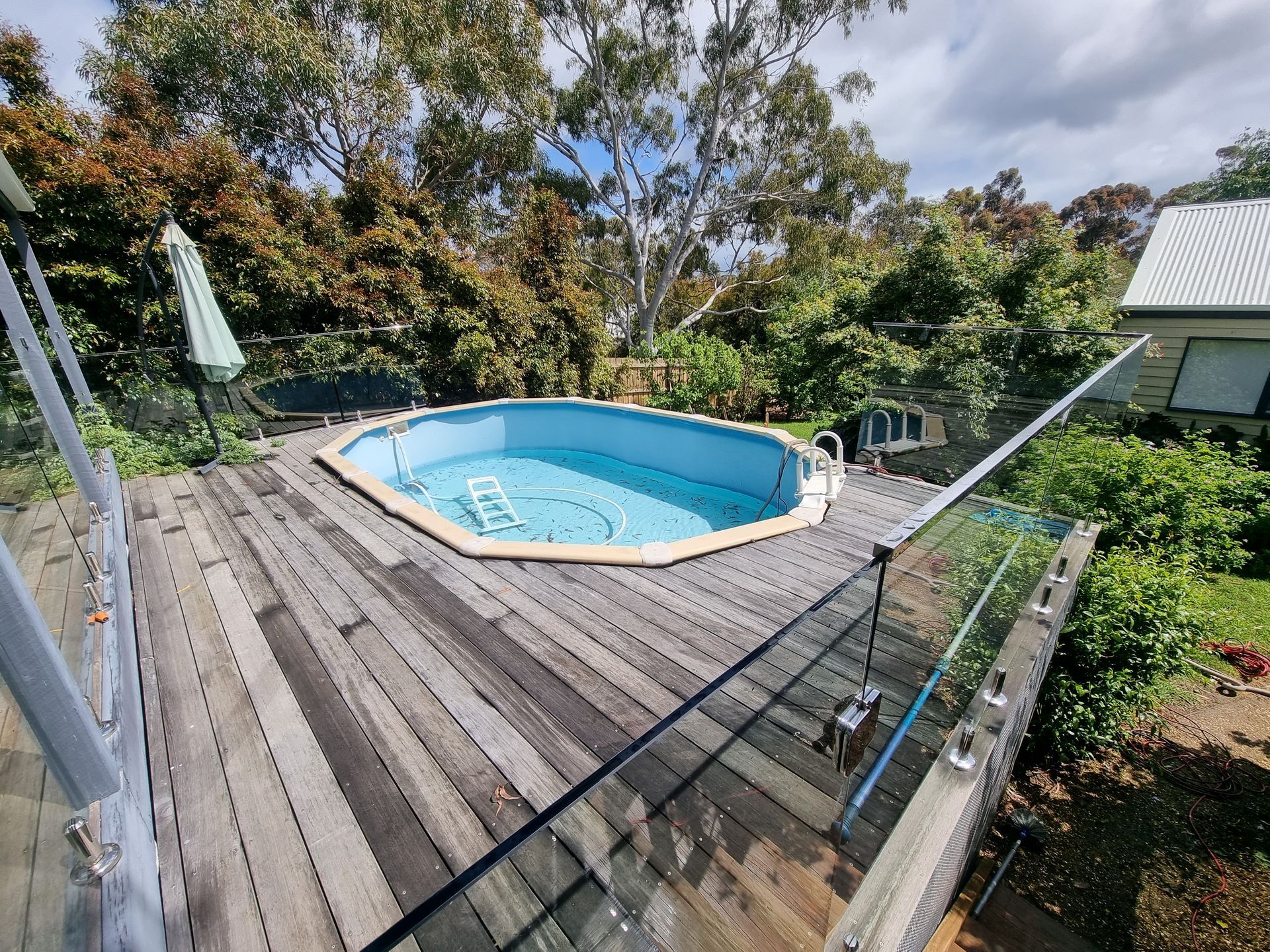 An octagonal above-ground swimming pool set into a wooden deck with glass railings, surrounded by trees — All Over Pool Removals in Chadstone, VIC