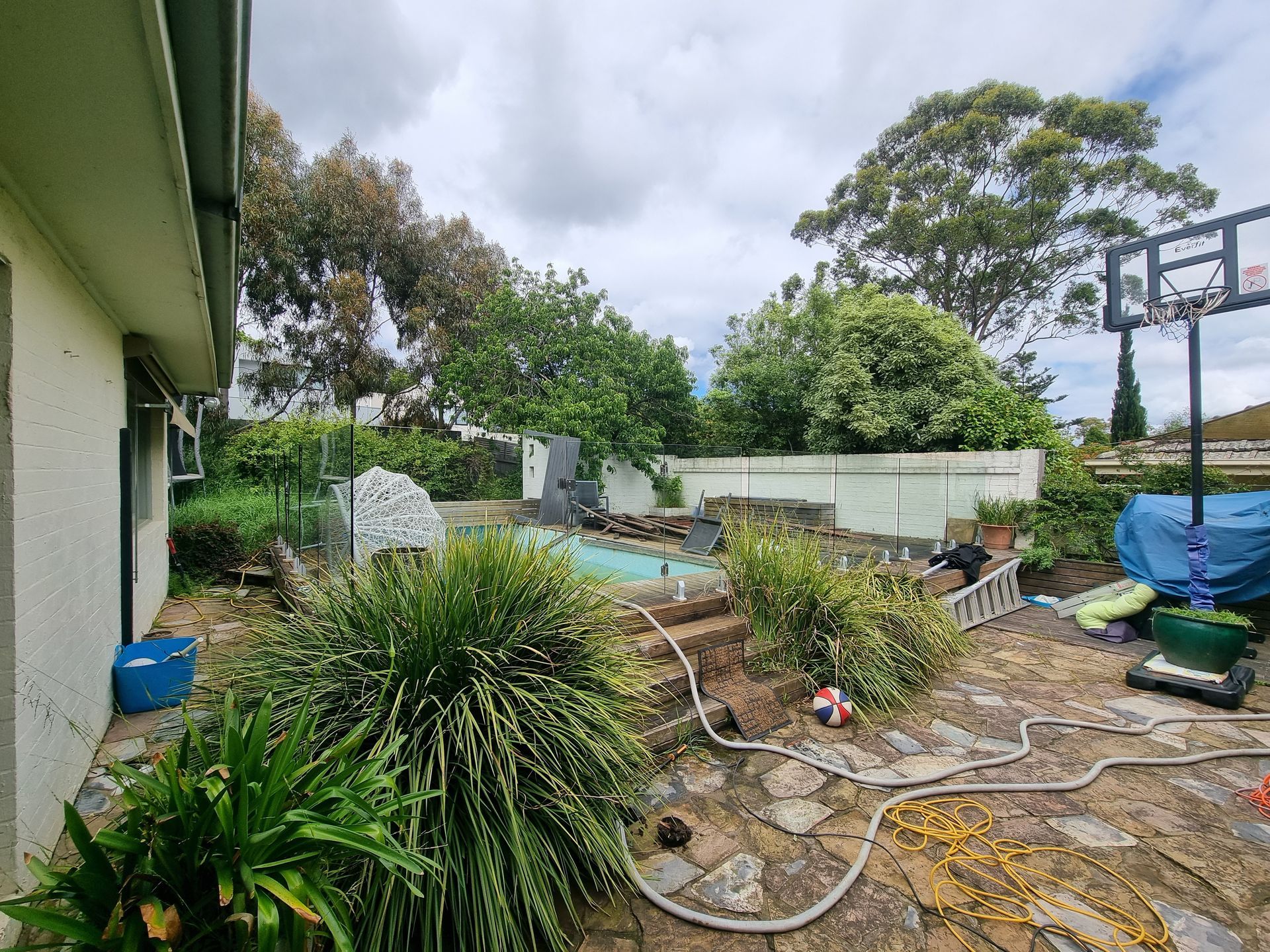A backyard with a swimming pool, stone patio, basketball hoop, and various plants under a cloudy sky  — All Over Pool Removals in Templestowe, VIC