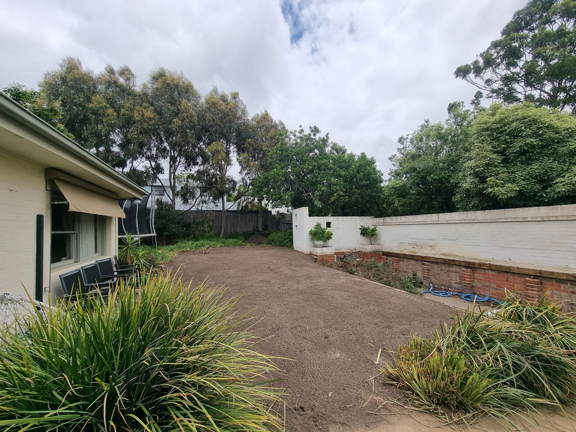 A backyard with a central area of loose brown soil, surrounded by garden beds, a beige house exterior, and a white wall — All Over Pool Removals in Hawthorn, VIC