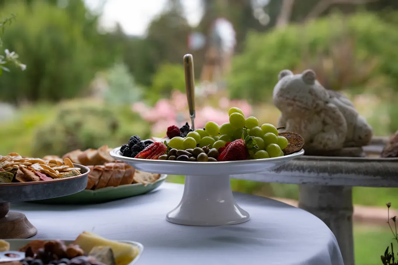 A fruit and cheese platter on a white cake stand outdoors on a table with a blurred garden background.