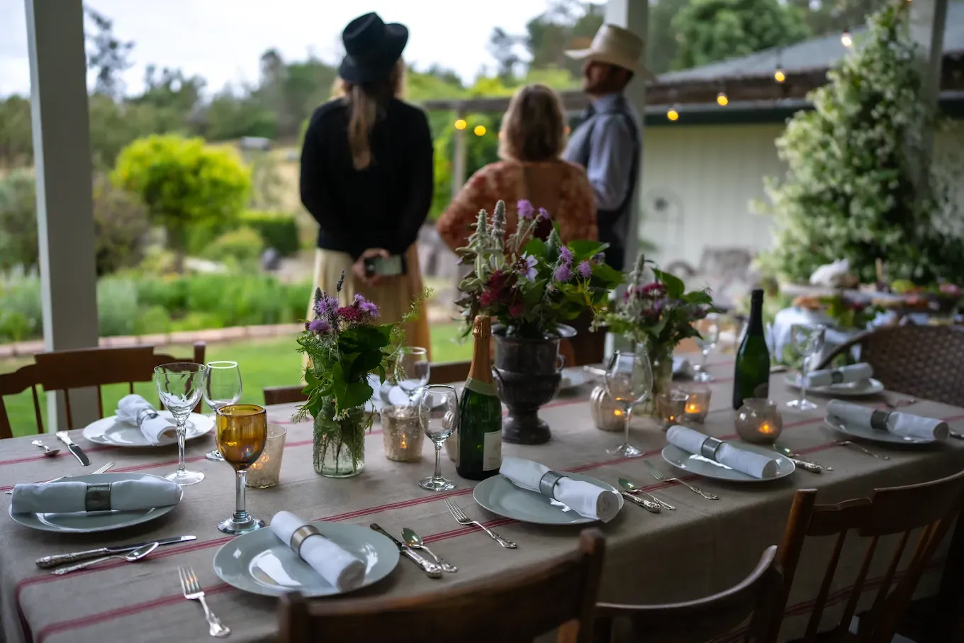 Elegant outdoor dining table set with flowers, people in background.