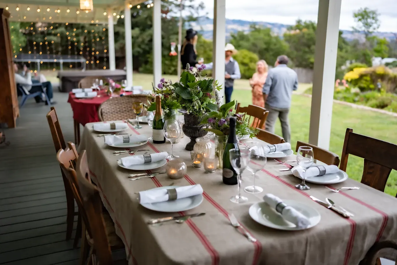 Long table set for a meal on a porch with guests standing nearby.