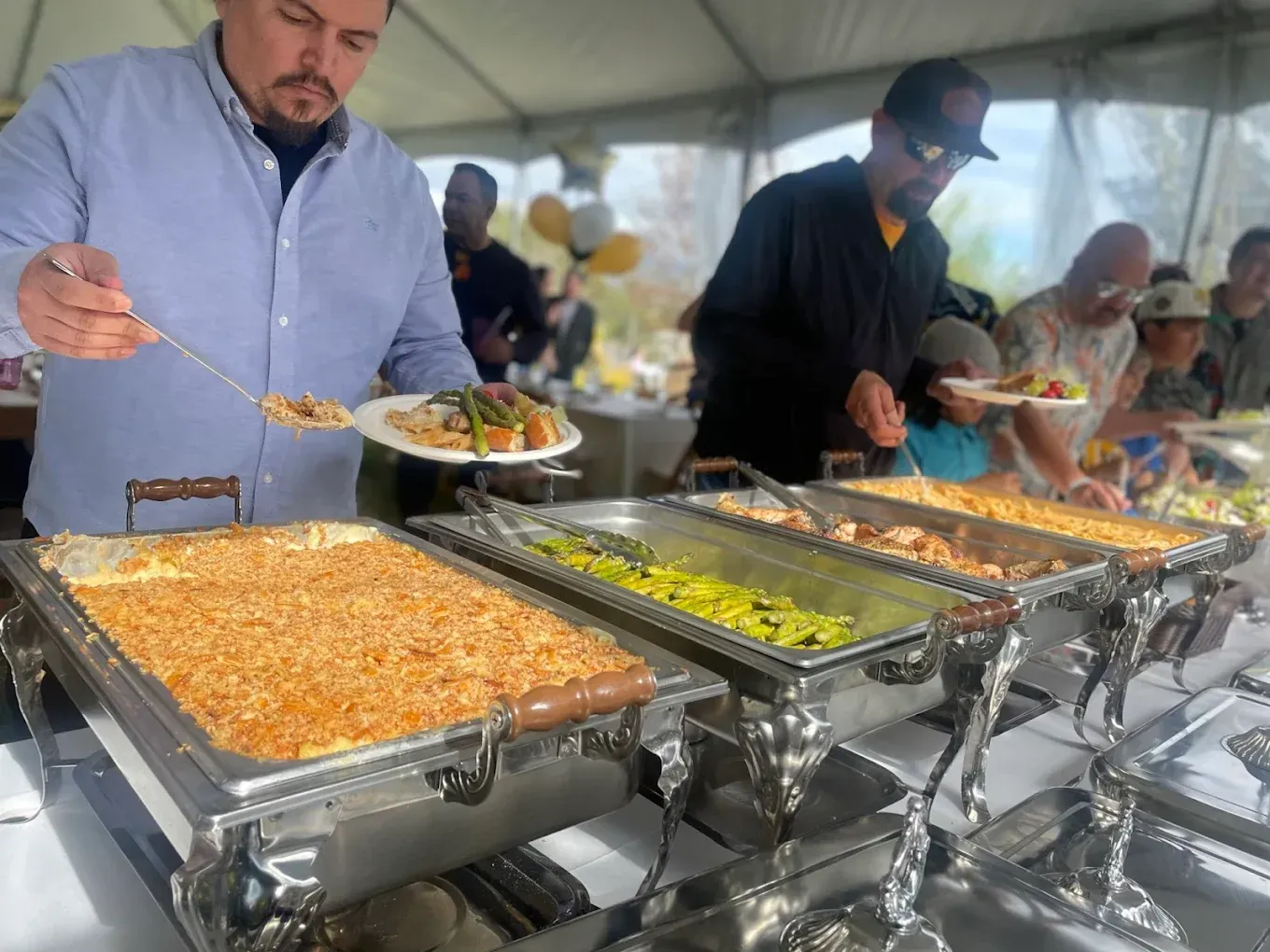 People at a buffet serving food from metal chafing dishes at an outdoor event.