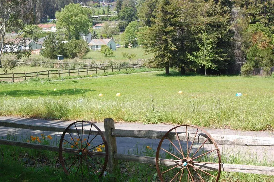 Grassy field with weathered fence and wagon wheels, houses in background. Sunny day.