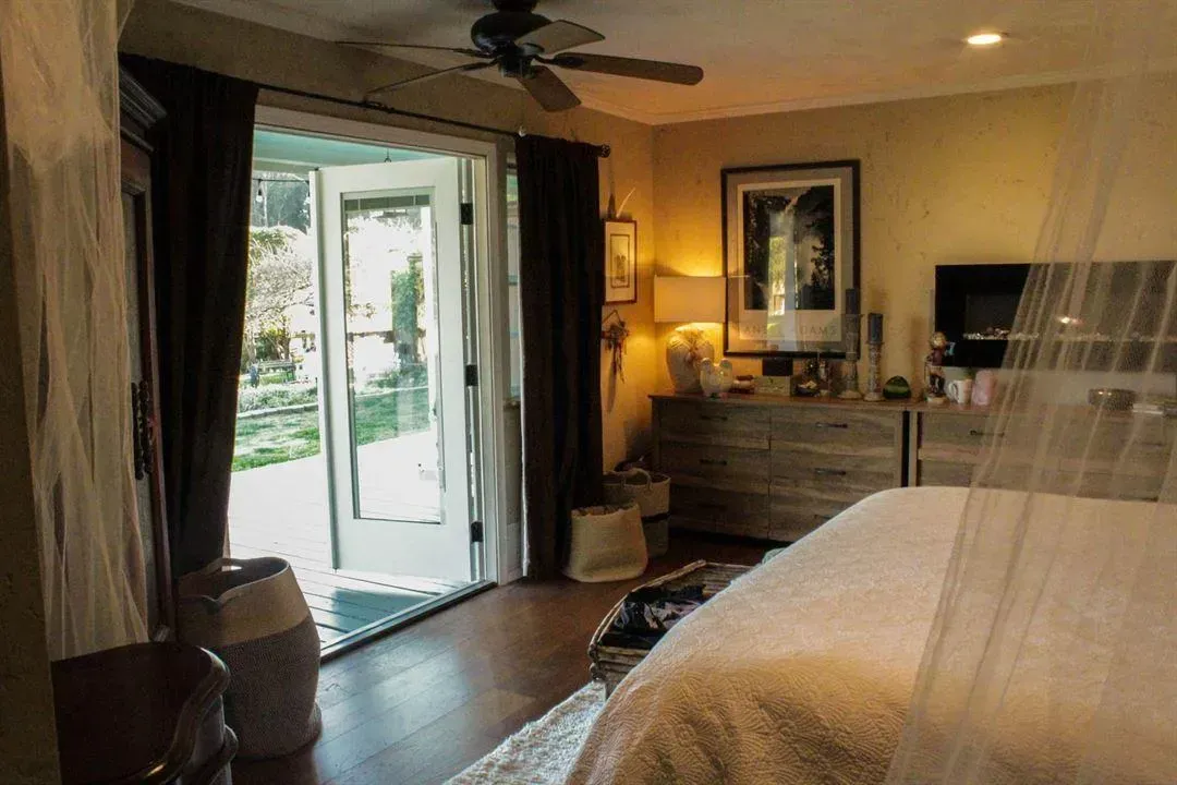 Bedroom with open doors to a yard. A bed, dresser, and dark curtains are visible.