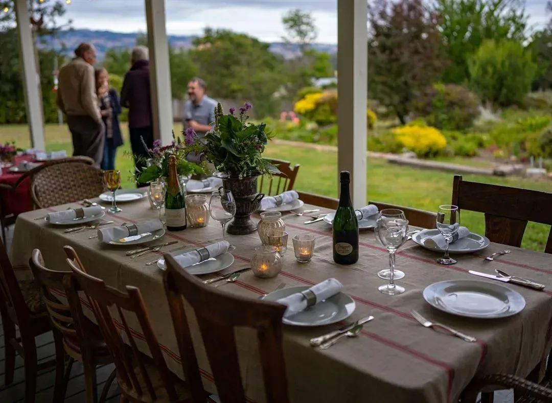 Outdoor dining table set for a meal; people in background on a porch with greenery.