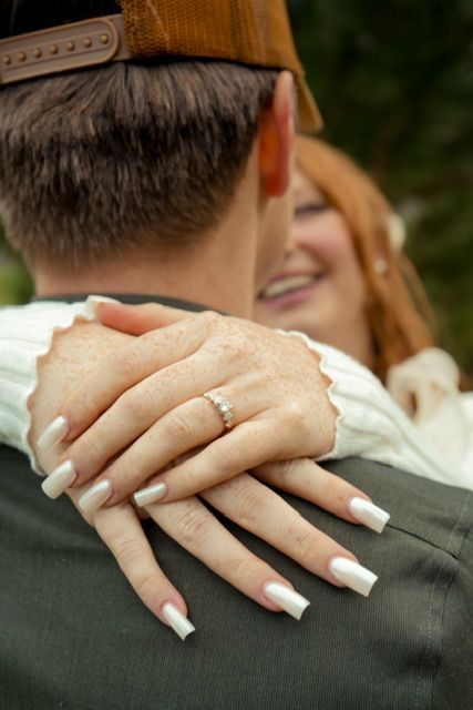 Woman's hands with white nails, wearing a ring, hugging a person with a brown hat. Smiling face visible.