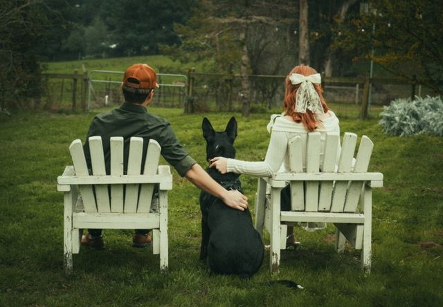 Couple in white chairs with black dog; man’s arm around dog, woman’s hand on dog, in a grassy yard.