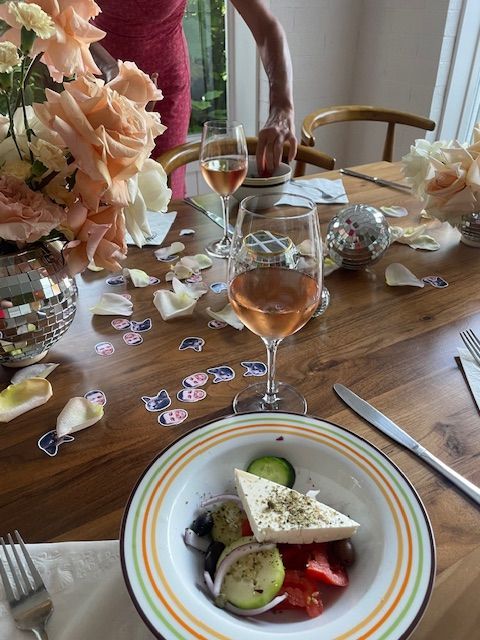 Greek salad and rosé on a wooden table with flowers and a disco ball; someone pours wine.