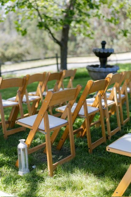 Wooden chairs with white cushions are arranged on grass near a fountain for an outdoor event.